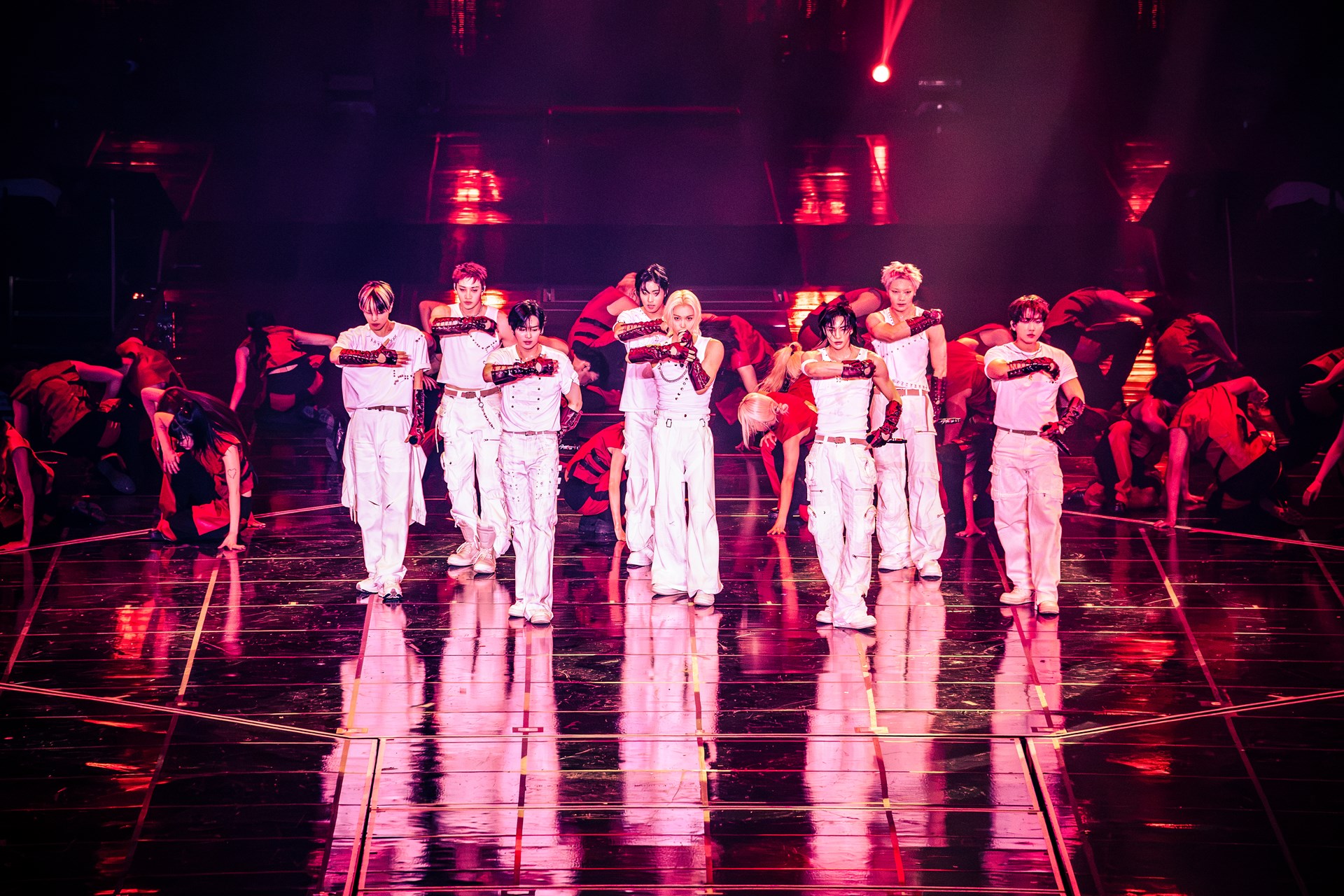 Eine K-Pop-Gruppe steht in weißen Outfits auf einer glänzenden Konzertbühne und führt eine synchronisierte Choreografie auf. Im Hintergrund tanzen weitere Tänzer. Rotes und pinkfarbenes Bühnenlicht taucht die Szene in eine intensive Atmosphäre und spiegelt sich auf dem dunklen Bühnenboden.